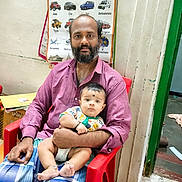 Viswa Guruparan is registered to the contest to win money with this photo: man, baby, child, indoor, red_chair, educational_poster, vehicles_poster, plaid_clothing, beard, casual_wear, floor, wall, doorway, curious_expression, holding, parent_and_child, portrait, seated, room, learning