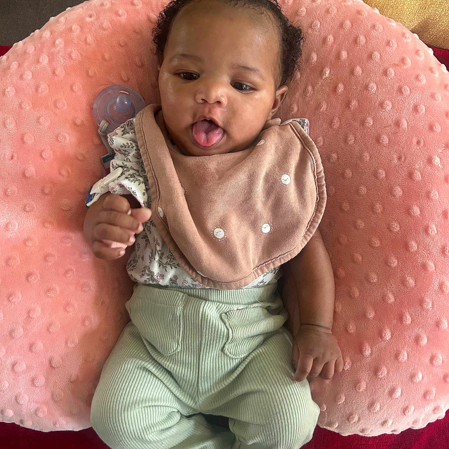Cimani is registered to the contest to win money with this photo: adorable, baby, bib, child, comfortable, cozy, cute, floral_top, flower_hair_clip, flower_socks, indoor, infant, lying_down, mint_green_pants, person, pink_pillow, playful, soft_texture, tongue_out, white_socks