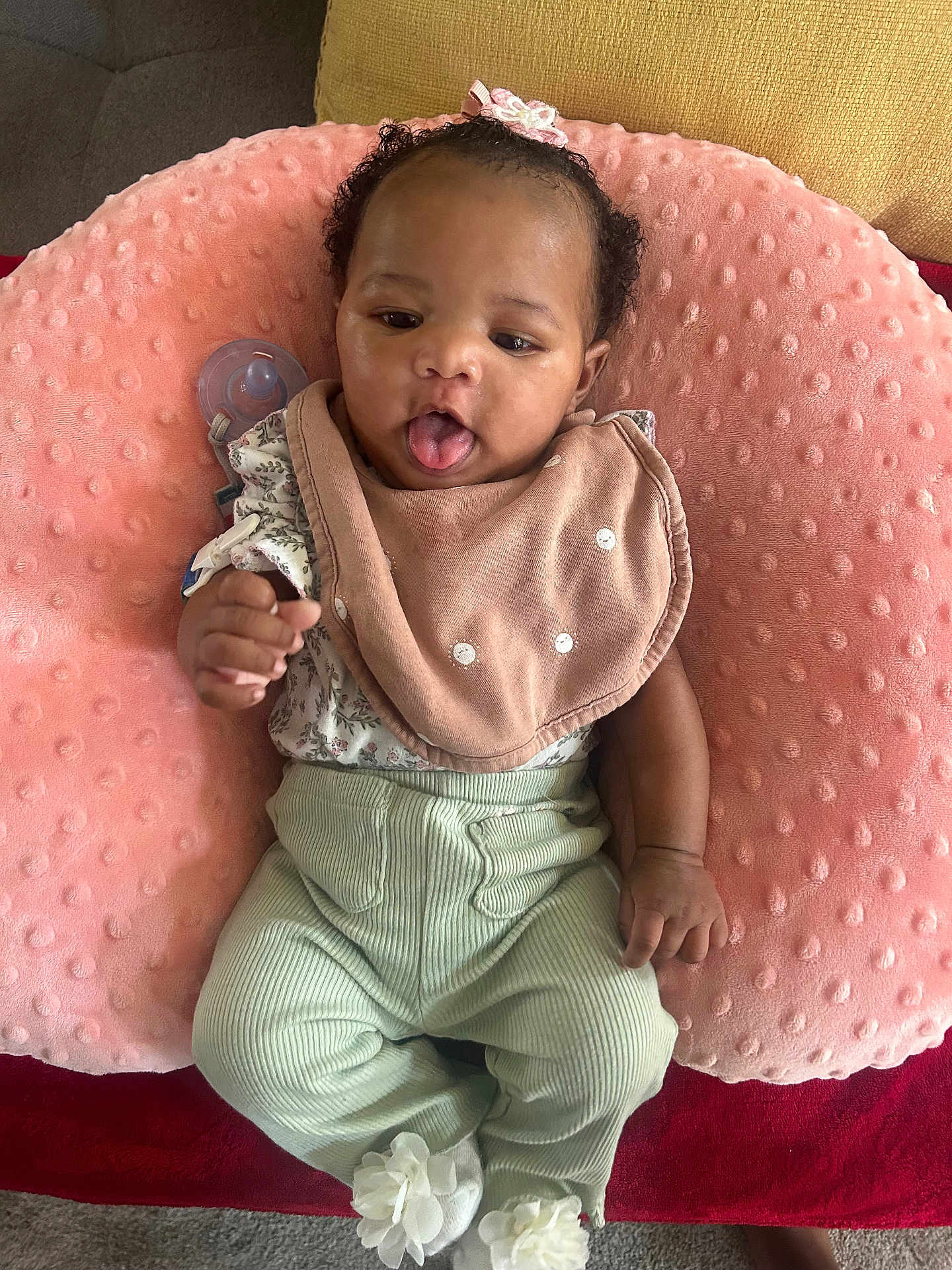 Cimani is registered to the contest to win money with this photo: baby, infant, pink_pillow, bib, flower_hair_clip, mint_green_pants, white_socks, flower_socks, tongue_out, cute, soft_texture, floral_top, lying_down, cozy, indoor, playful, child, person, adorable, comfortable