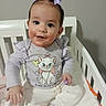 baby, child, crib, purple_bow, cartoon_cat, shirt, white_pants, socks, indoor, happy, smiling, hand, face, hair, person, furniture, bed, cute, infant, home