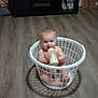 baby, infant, laundry_basket, bottle, floor, wooden_floor, smiling, indoors, television, cabinet, furniture, milk, child, cute, sitting, home, room, object, happy, person