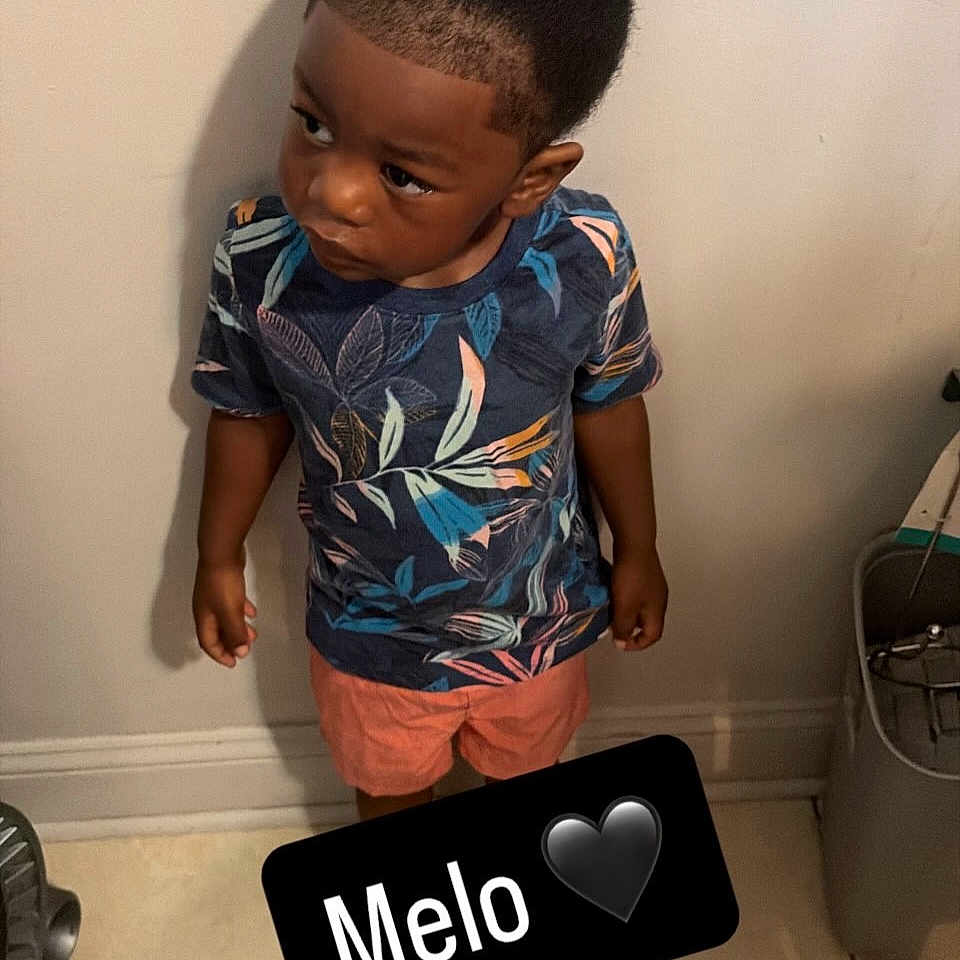 Carmelo joined the competition — help win amazing prizes! child, boy, standing, indoor, tiled_floor, shorts, tshirt, patterned_clothing, hairstyle, curious_expression, wall, floor, shadow, household_item, black_text_overlay, emoji, portrait, casual_clothing, young, person