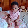 baby, infant, child, pink_bib, blanket, stuffed_animal, teddy_bear, wood_paneling, bed, curly_hair, wide_eyes, sitting, indoors, cute, portrait, clothing, soft_toys, furniture, skin, person