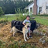 child, boy, dog, german_shepherd, outdoor, portrait, standing, smiling, pet, animal, grass, weeds, tire, garden, yard, house, rural, chimney, weathered, daylight