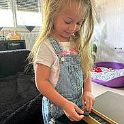 Kelya a rejoint le concours — aidez-le/la à gagner de superbes lots ! child, girl, blonde_hair, denim_overalls, white_tshirt, playing, wooden_box, indoor, window, couch, pet_bed, carpet, furniture, smiling, happy, casual_clothing, long_hair, person, home, daylight