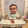 baby, high_chair, bib, pacifier, table, toy, indoor, child, face, smile, curious, furniture, wooden_table, wallpaper, window_blinds, home, infant, clothing, person, cute