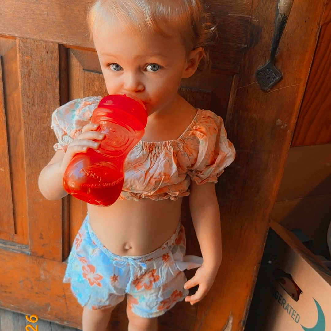 Coralyn is registered to the contest to win money with this photo: toddler, child, barefoot, red_sippy_cup, floral_clothing, wooden_floor, wooden_door, pony_tail, indoor, standing, cute, drink, box, vintage_filter, date_stamp, door_handle, light, person, young_child, casual_clothing