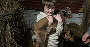 Abal is registered to the contest to win money with this photo: child, baby_goat, barn, hay, animal, farm, rustic, brown_clothing, boots, smile, holding, livestock, indoor, person, young, goat, fence, wood, metal_sheets, dark