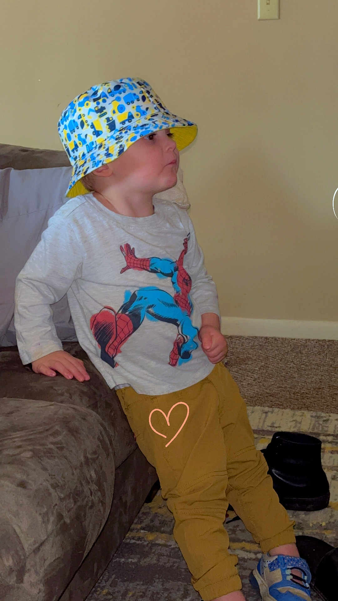 Carlisle is registered to the contest to win money with this photo: toddler, child, bucket_hat, spiderman_shirt, yellow_pants, blue_sneakers, couch, carpet, indoor, wall, footwear, person, side_view, casual_pose, expression, young_child, clothing, furniture, home, leaning