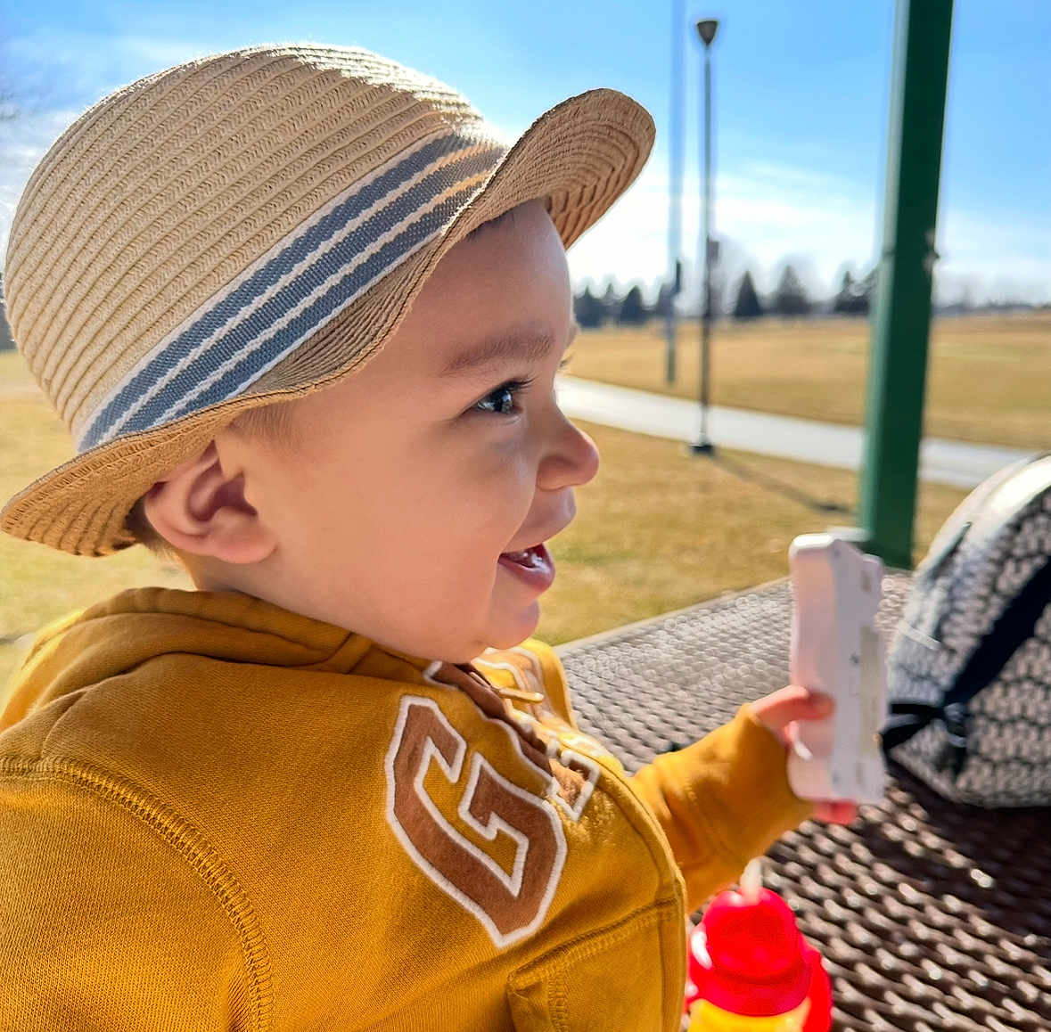 Matthew joined the competition — help win amazing prizes! toddler, child, hat, straw_hat, yellow_hoodie, smiling, side_profile, outdoor, park, picnic_table, toy, daylight, happy, face, person, sunny, background_blur, hand, sippy_cup, casual_clothing