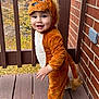 toddler, child, costume, lion_costume, smiling, outdoor, porch, wooden_floor, brick_wall, autumn, leaves, cute, baby, face, person, standing, happy, animal_costume, footwear, tail