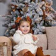 Idalia joined the competition — help win amazing prizes! child, toddler, smiling, curly_hair, hair_bows, sweater, brown_socks, couch, christmas_tree, ornaments, snow_flocked, lights, gift_box, pinecone, holiday, indoor, festive, decorations, happy, portrait