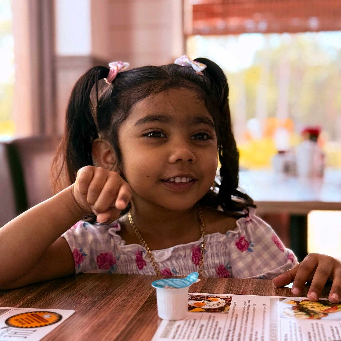 Ariella joined the competition — help win amazing prizes! blurred_background, casual_clothing, child, creamer_cup, cute, daylight, food, girl, happy, indoor, jewelry, menu, person, pigtails, playful, portrait, restaurant, smiling, table, wooden_table