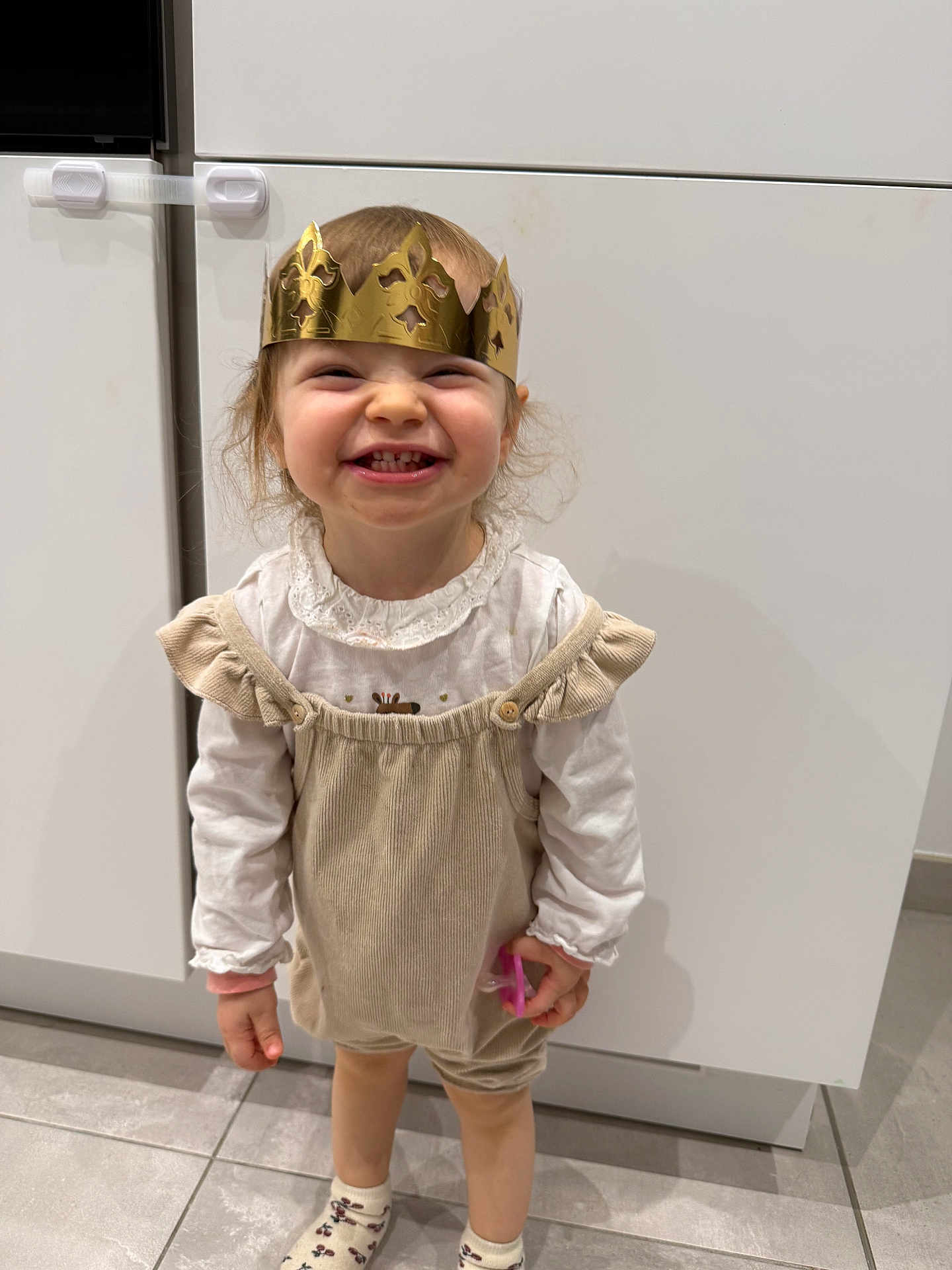 Elif a rejoint le concours — aidez-le/la à gagner de superbes lots ! toddler, child, smiling, crown, gold, indoor, white_clothing, beige_romper, floor_tiles, cabinet, happy, cute, small_object, playful, person, young_child, standing, long_sleeve, sock, portrait