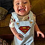 baby, child, smiling, superman, overalls, clothing, indoor, couch, happy, tongue_out, person, carpet, bag, cute, playful, shorts, footwear, bright, portrait, young