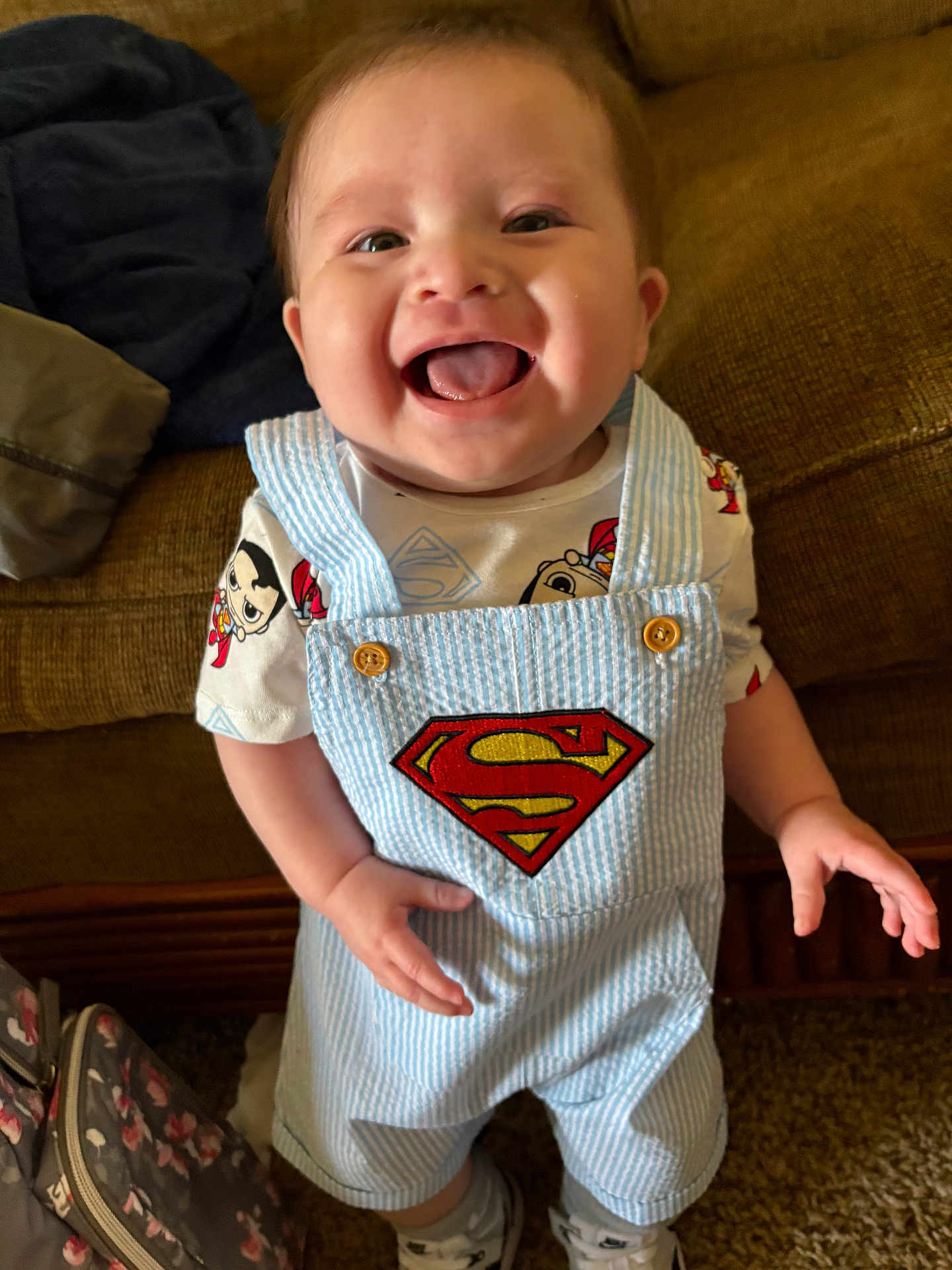 Ezequiel joined the competition — help win amazing prizes! baby, child, smiling, superman, overalls, clothing, indoor, couch, happy, tongue_out, person, carpet, bag, cute, playful, shorts, footwear, bright, portrait, young