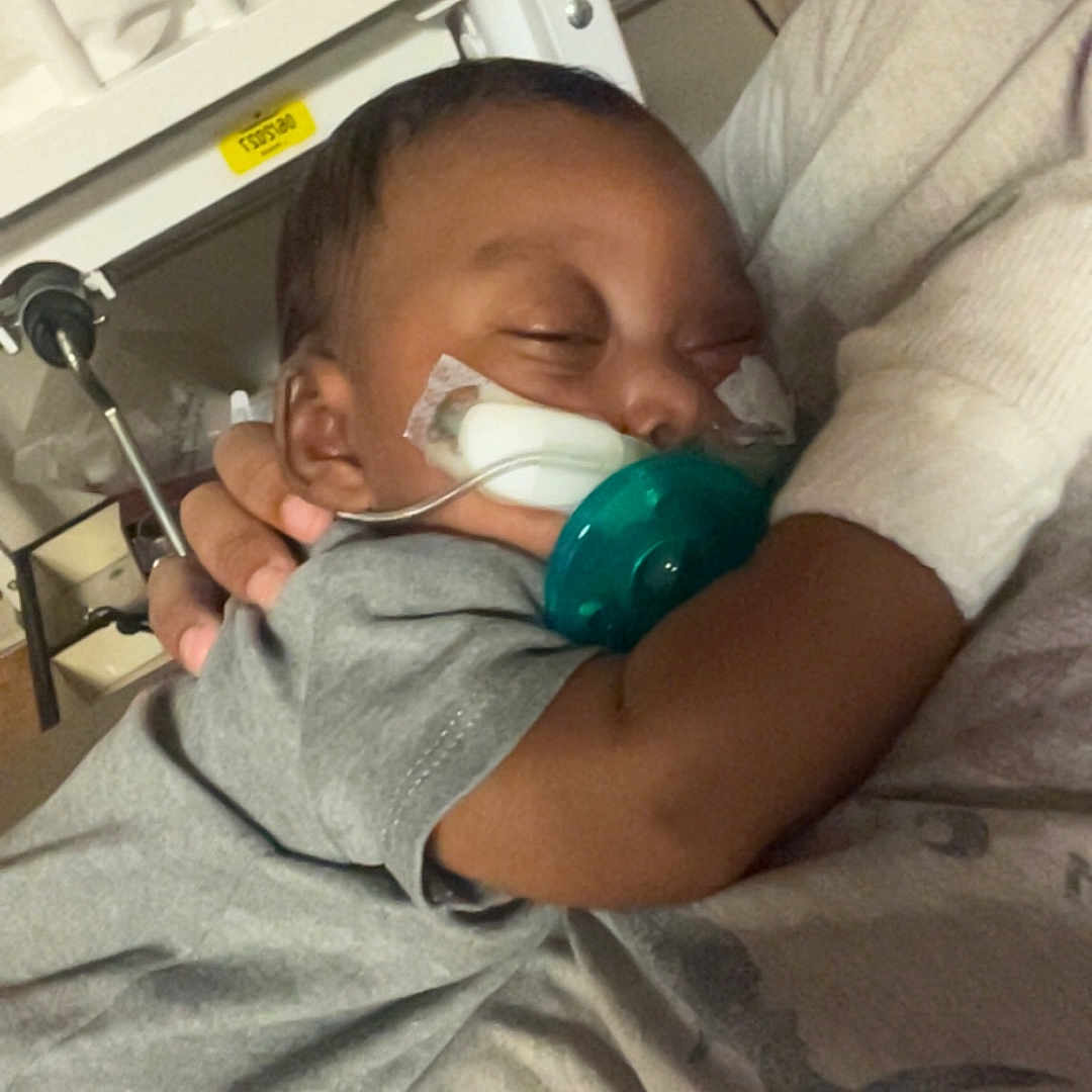 Hezekiah joined the competition — help win amazing prizes! adult, baby, bed, bed_rail, blanket, caregiving, closed_eyes, cuddling, face, gray_clothing, hospital, indoors, infant, medical_tubing, pacifier, parent, photograph, portrait, resting, sleeping