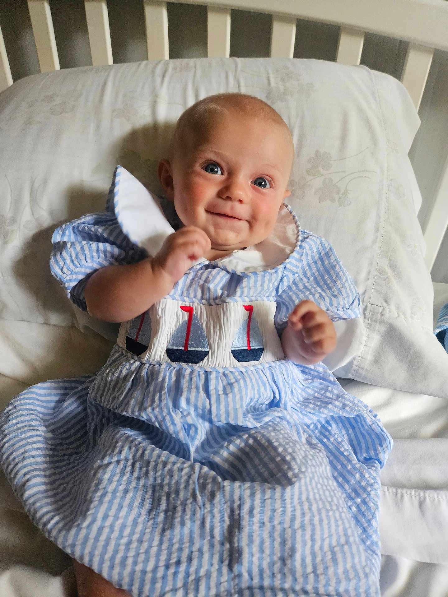 Atillia joined the competition — help win amazing prizes! baby, child, blue_dress, striped_dress, sailboat_embroidery, smiling, sitting, bed, pillow, crib, indoor, person, portrait, happy, infant, cute, clothing, face, head, human