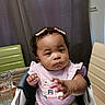 baby, infant, child, high_chair, sitting, indoor, portrait, face, curly_hair, hair_bow, pink_clothing, bib, reaching, hand, curious, neutral_expression, furniture, curtains, home, photograph