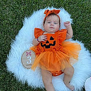 Victoria joined the competition — help win amazing prizes! baby, infant, child, halloween, pumpkin_costume, orange_dress, bow, grass, outdoor, milestone_photo, three_months, fluffy_blanket, jack_o_lantern, lying_down, portrait, cute, baby_photography, raised_fist, wooden_sign, tulle_skirt