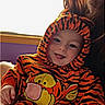 Cory is registered to the contest to win money with this photo: baby, child, tiger_onesie, orange_clothing, smiling, happy, indoor, window, adult, person, face, hooded_clothing, toddler, cute, cozy, purple_wall, bright_light, holding, tongue_out, casual