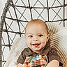 baby, child, smiling, dimple, plaid_overalls, brown_shirt, hanging_chair, woven_chair, blanket, cozy, indoors, portrait, cute, happy, playful, infant, person, seated, bright, soft_light