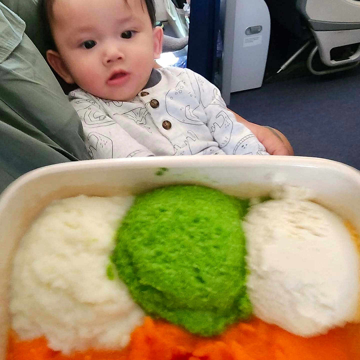 Ryu Nguyen joined the competition — help win amazing prizes! airplane, baby, blurred_background, buttoned_shirt, child, clothing, curious, face, food, green_mash, hand, holding, indoor, mashed_food, orange_mash, passenger, person, seat, tray, white_mash