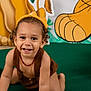 toddler, child, smiling, crawling, brown_clothing, yellow_hat, beads_necklace, green_carpet, balloons, jungle_theme, decorations, curly_hair, happy, playful, portrait, indoors, cute, baby, person, fun