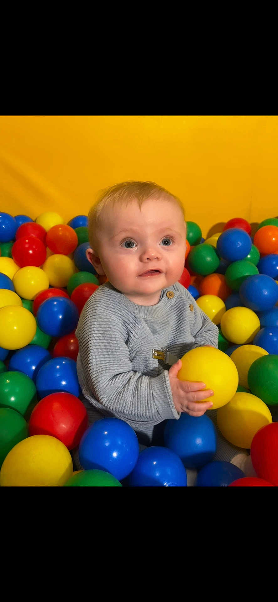 Finley is registered to the contest to win money with this photo: baby, child, playing, ball_pit, colorful, plastic_balls, yellow, blue, red, green, orange, sweater, indoor, cute, smiling, happy, portrait, young, fun, play