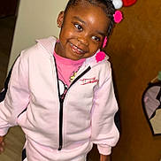 Lamiyah is registered to the contest to win money with this photo: child, girl, smile, happy, indoor, tracksuit, pink_clothing, hairstyle, accessories, earrings, wooden_door, floor, person, portrait, fashion, casual_wear, cute, standing, young_child, joy