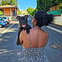 Arlow participe au concours pour gagner de l'argent avec cette photo : back_view, black_dog, building, car, daytime, dog, fence, floral_dress, happy, holding, leisure, nature, outdoor, pavement, person, sidewalk, smiling_dog, street, sunny, trees