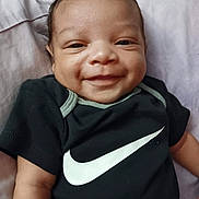 Carmelo is registered to the contest to win money with this photo: baby, infant, smiling, face, eyes, nose, mouth, onesie, nike, logo, black_shirt, arm, blanket, pillow, lying_down, portrait, close_up, happy, hair, skin_tone