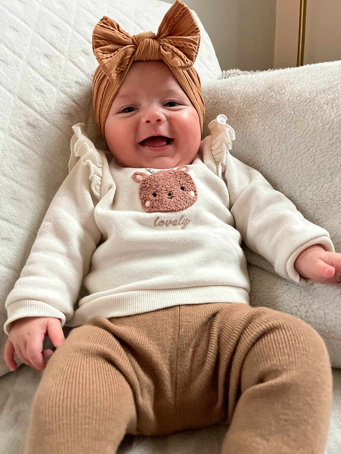 Anaya participe au concours pour gagner de l'argent avec cette photo : baby, infant, smiling, headband, bow, blanket, sweater, embroidered_patch, bear_motif, neutral_tones, cozy, couch, plush, portrait, indoor, clothing, hand, legs, knit_pants, happy