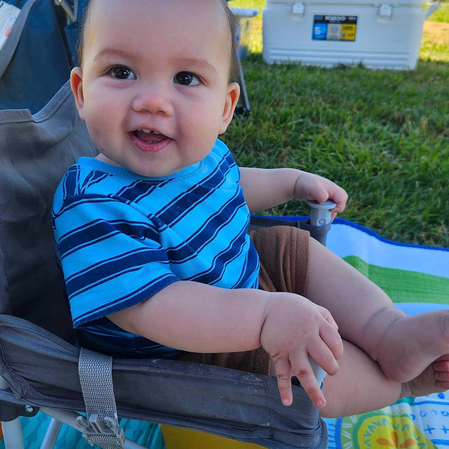 Jax joined the competition — help win amazing prizes! baby, child, smiling, blue_shirt, striped_shirt, outdoor, grass, chair, play_mat, happy, face, person, cute, summer, daylight, nature, legs, hand, seat, infant