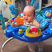 Jax joined the competition — help win amazing prizes! baby, child, activity_jumper, toy, orange_star, red_crab, blue, carpet, bookshelf, books, indoor, floor, clothing, socks, play, smile, person, infant, happy, colorful