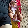 Aleena is registered to the contest to win money with this photo: baby, infant, pink_headband, bib, hammock, adult_arm, wooden_floor, black_clothing, baby_shoes, pink_shoes, outdoor, person, relaxing, colorful_hammock, cute, expression, casual, summer, resting, portrait