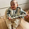 baby, child, smiling, indoor, plaid_jacket, knitwear, wooden_floor, cozy_chair, holding_object, decorative_sign, happy, cute, portrait, person, home_interior, soft_light, flooring, socks, seated, infant
