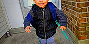 Kyson is registered to the contest to win money with this photo: toddler, child, smiling, walking, backpack, puffer_vest, sneakers, concrete, porch, door, brick_wall, curly_hair, outdoor, casual_clothing, happy, person, young_child, sidewalk, urban, daylight