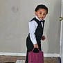 Kyson is registered to the contest to win money with this photo: child, boy, indoor, formal_wear, vest, bow_tie, black_pants, white_shirt, purple_bag, curly_hair, standing, floor, door, carpet, paper, shoe, person, portrait, young, curious_expression