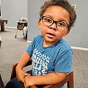 Kyson is registered to the contest to win money with this photo: child, glasses, curly_hair, blue_shirt, wooden_chair, indoor, carpet, high_chair, pool_table, person, seated, young, casual_clothing, furniture, curious, portrait, face, eyewear, expression, home