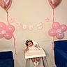 baby, high_chair, balloon, pink, heart_balloon, flower_balloon, banner, celebration, birthday, smiling, child, indoor, headband, dress, wooden_floor, blue_chair, wall, happy, person, toddler