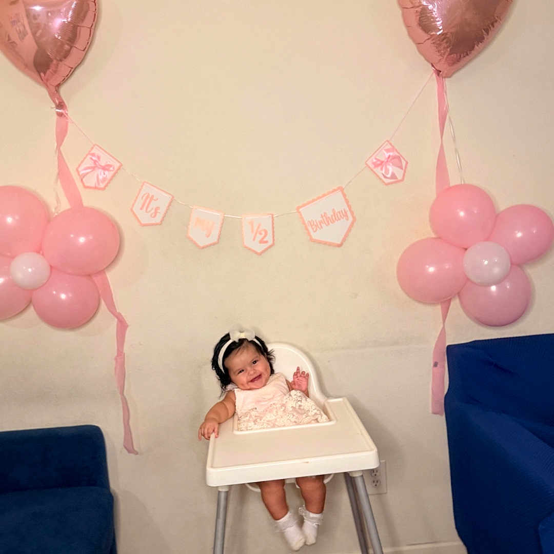 Keilany joined the competition — help win amazing prizes! baby, high_chair, balloon, pink, heart_balloon, flower_balloon, banner, celebration, birthday, smiling, child, indoor, headband, dress, wooden_floor, blue_chair, wall, happy, person, toddler