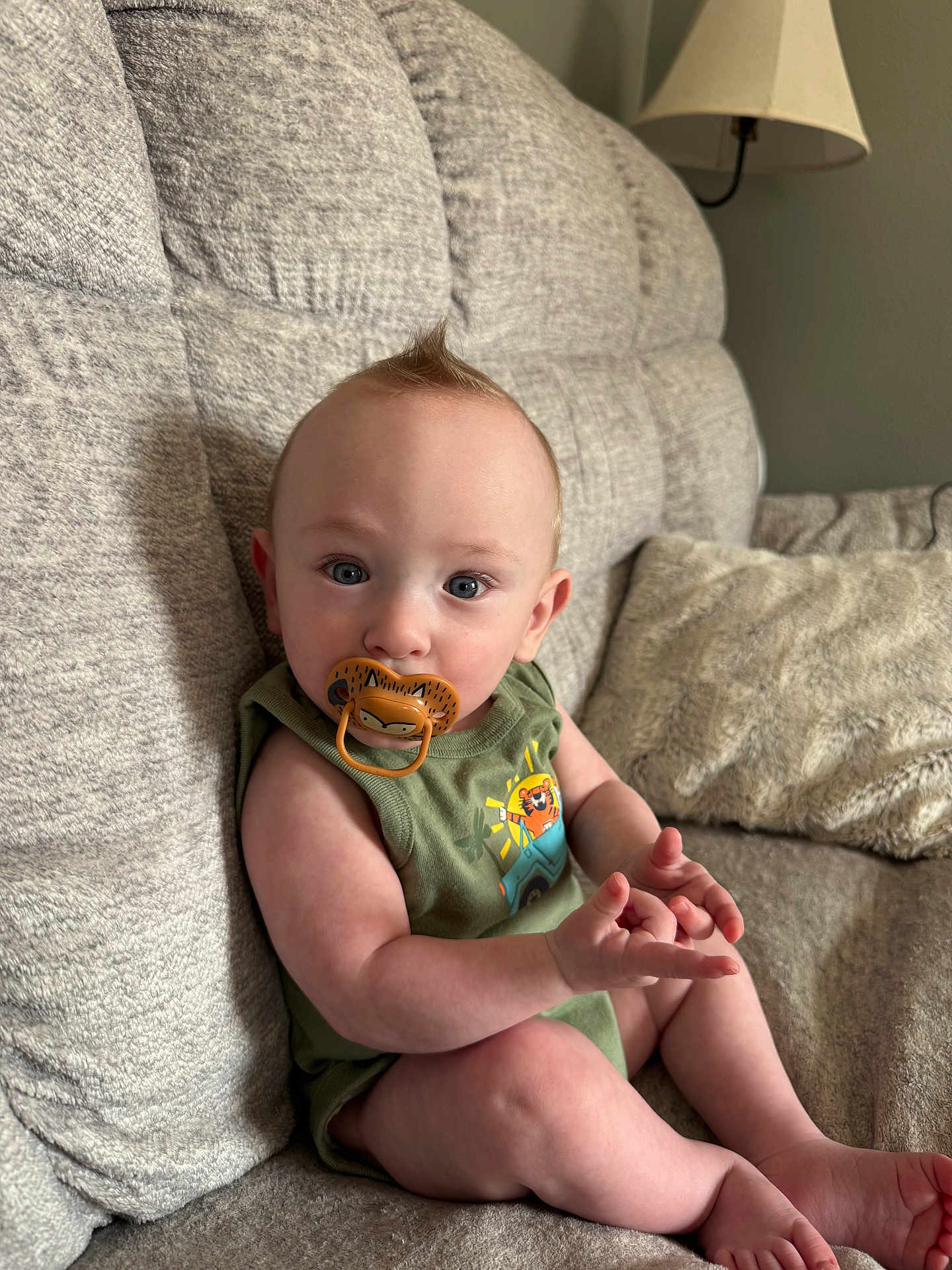 Jude is registered to the contest to win money with this photo: baby, pacifier, green_clothing, couch, gray_couch, indoor, child, infant, blue_eyes, sitting, plush_pillow, lamp, mohawk_hairstyle, cute, toddler, portrait, furniture, home_interior, person, relaxed