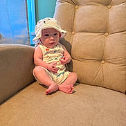 Jude is registered to the contest to win money with this photo: baby, child, hat, white_hat, bear_face, sitting, curious, beige_chair, cushion, indoor, window, natural_light, skin, feet, arms, clothing, infant, cute, face, portrait
