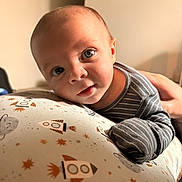 Grayson is registered to the contest to win money with this photo: baby, infant, face, blue_eyes, clothing, striped_shirt, hand, cushion, space_theme, rocket, stars, planet, indoor, portrait, child, resting, curious, smile, person, closeup