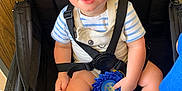 Ozzee is registered to the contest to win money with this photo: toddler, child, blue_ribbon, stroller, curly_hair, striped_shirt, overalls, shoes, indoor, award, seatbelt, hand, face, expression, toy, paper, black_stroller, light, furniture, person