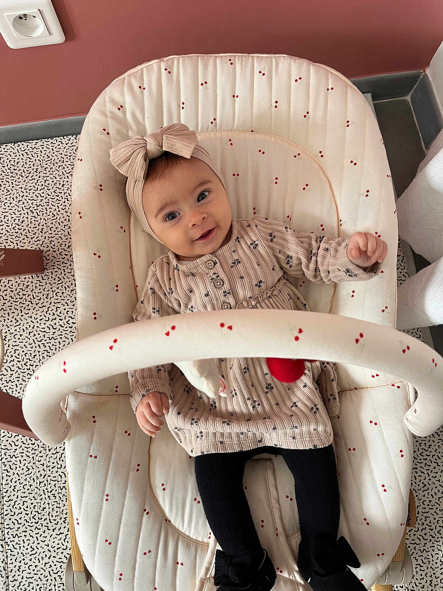 Lucy a rejoint le concours — aidez-le/la à gagner de superbes lots ! baby, child, infant, headband, bow, dress, patterned_clothing, black_tights, baby_seat, cushion, cherry_pattern, smiling, happy, indoors, floor_pattern, pink_wall, power_socket, furniture, toy_bar, portrait