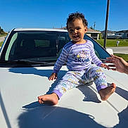 Hannah is registered to the contest to win money with this photo: toddler, child, car, white_car, pajamas, purple_clothing, smiling, outdoor, daylight, shadow, hand, curly_hair, hood, street, residential_area, blue_sky, sunny, sitting, playful, happy
