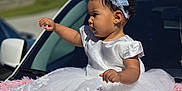 Hannah is registered to the contest to win money with this photo: baby, child, dress, white_dress, headband, pink_blanket, car, outdoor, curious, sitting, portrait, sunny, cute, infant, fashion, accessory, vehicle, background, daylight, expression