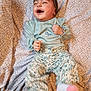 baby, child, laughing, headband, green_clothing, floral_pants, socks, patterned_bedspread, happy, infant, smiling, cute, toddler, indoors, portrait, person, clothing, fabric, bed, playful